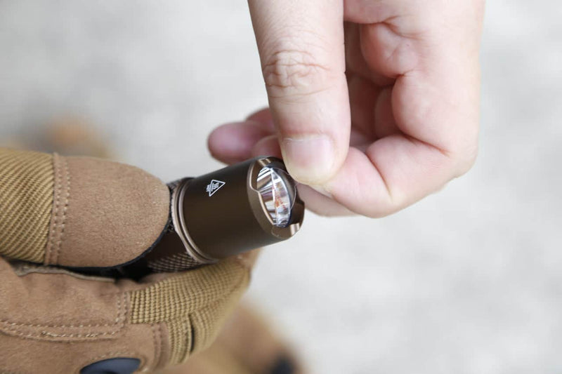 A person uses their thumb and forefinger to peel protective film from the lens of a Manker MC11 III USB-C Rechargeable Tactical Flashlight held in a gloved hand.