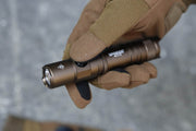 A person wearing brown tactical gloves holds a Manker MC11 III USB-C Rechargeable Tactical Flashlight outdoors.