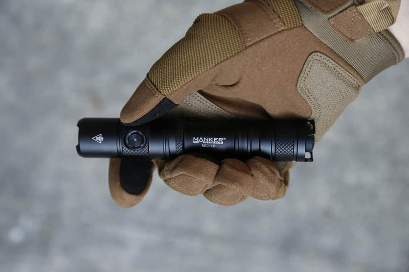 A gloved hand holds a black Manker MC11 III USB-C Rechargeable Tactical Flashlight against a gray background.