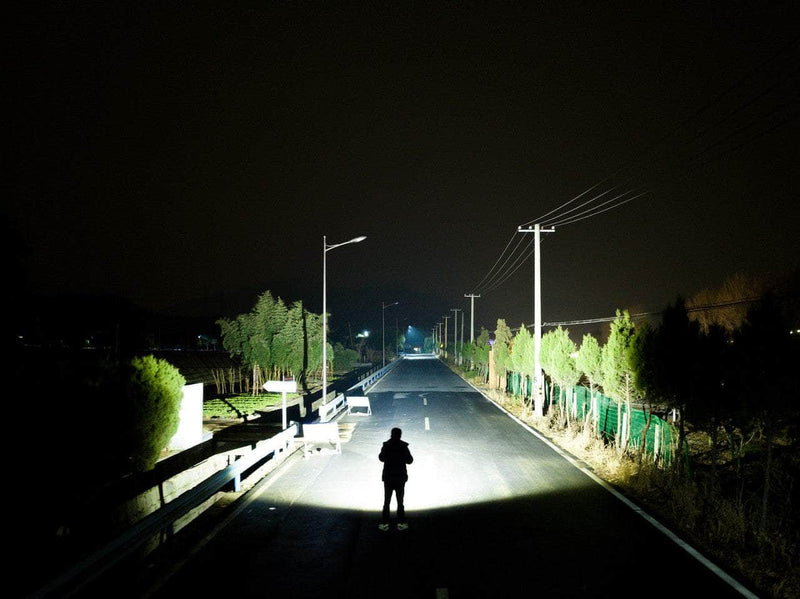 Under the bright illumination of the Manker Ranger MK39 II, a person stands at night in the middle of a road flanked by trees, with power lines running overhead.