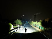 A person stands in the middle of an empty road at night, illuminated by the bright Manker Ranger MK39 II spotlight. The road is flanked by trees and power lines.