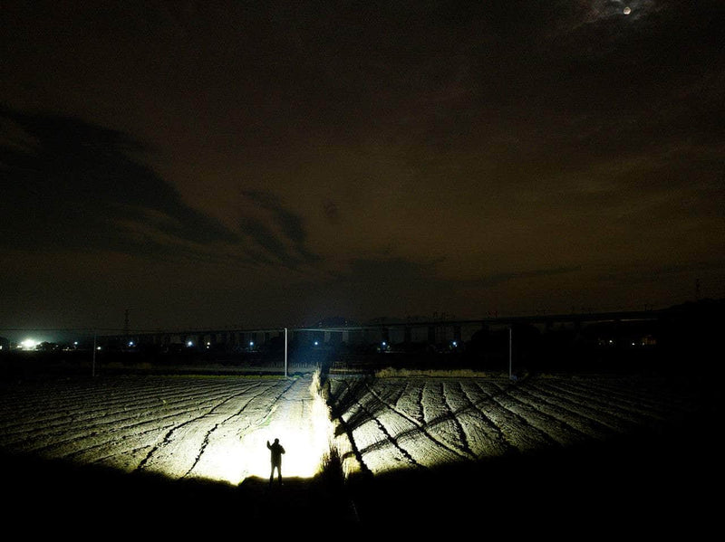 The Manker Ranger MK39 II casts a powerful beam on a figure standing in the field at night, silhouetted against a dark sky with scattered clouds.