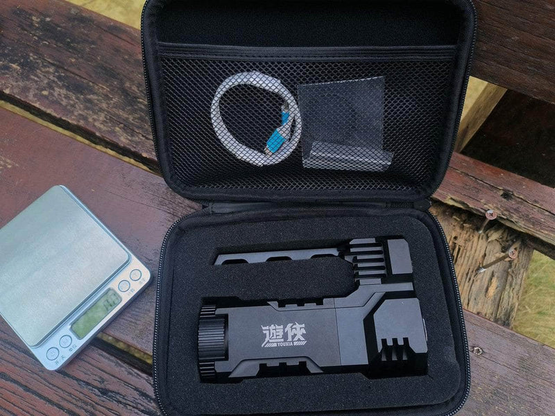 Open a black case containing a compact tool, USB cable, and small netted pouch. Beside it on the wooden surface sits a digital scale, all illuminated by the soft glow of the Manker Ranger MK39 II long-range spotlight.