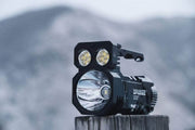 A Manker Ranger MK39 II, a black flashlight with multiple LED bulbs and spotlight feature, rests on a wooden post against a blurred mountainous background.