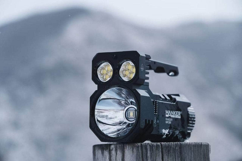 A Manker Ranger MK39 II, a black flashlight with multiple LED bulbs and spotlight feature, rests on a wooden post against a blurred mountainous background.
