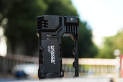 The Manker Ranger MK39 II flashlight, with its sleek angular design and powerful spotlight and floodlight combo, stands outdoors against a blurred backdrop of trees and buildings.