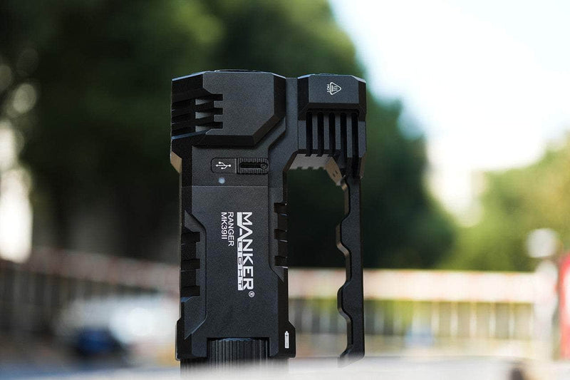 The Manker Ranger MK39 II flashlight, with its sleek angular design and powerful spotlight and floodlight combo, stands outdoors against a blurred backdrop of trees and buildings.