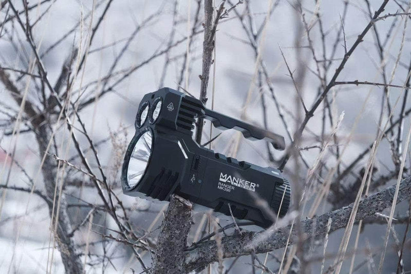 The Manker Ranger MK39 II flashlight, resembling a spotlight, rests on tree branches in a snowy landscape.