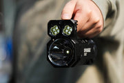 A close-up captures someone holding the Manker Ranger MK39 II flashlight, which features dual square lens clusters alongside a larger round lens designed for long-range spotlight.