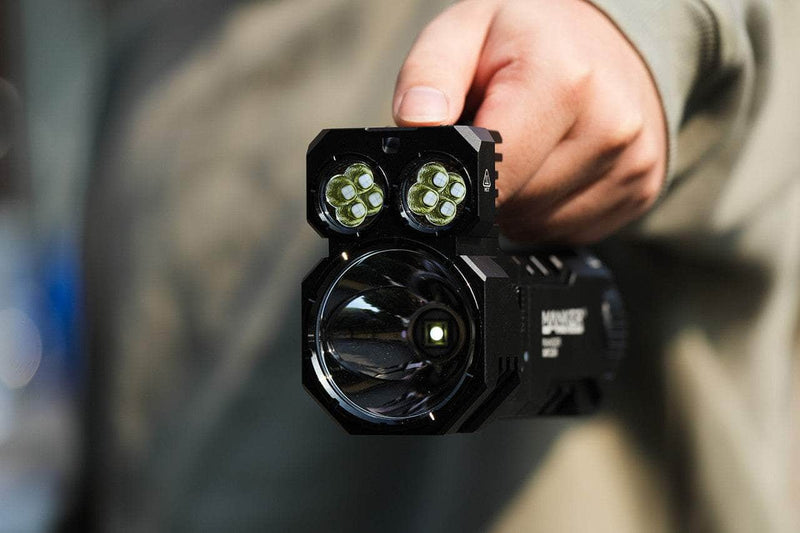 A close-up captures someone holding the Manker Ranger MK39 II flashlight, which features dual square lens clusters alongside a larger round lens designed for long-range spotlight.