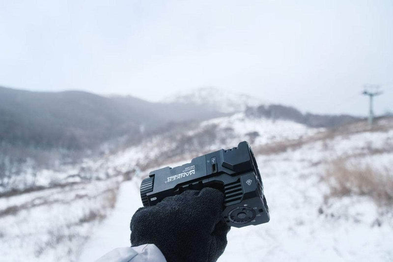A gloved hand holds the sleek, black Manker Ranger MK39 II against a snowy mountain landscape under a cloudy sky, casting a spotlight on the breathtaking vista.