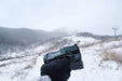 A gloved hand holds the sleek, black Manker Ranger MK39 II against a snowy mountain landscape under a cloudy sky, casting a spotlight on the breathtaking vista.
