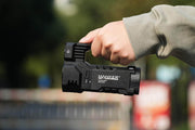 A hand grips the sleek black Manker Ranger MK39 II flashlight outdoors, with its powerful spotlight piercing through the blurred background of trees and a red sign.