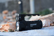 A Manker Ranger MK39 II flashlight, similar to a long-range spotlight, is set outdoors on a concrete surface surrounded by broken bricks and scattered debris.