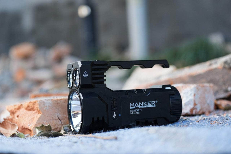 A Manker Ranger MK39 II flashlight, similar to a long-range spotlight, is set outdoors on a concrete surface surrounded by broken bricks and scattered debris.