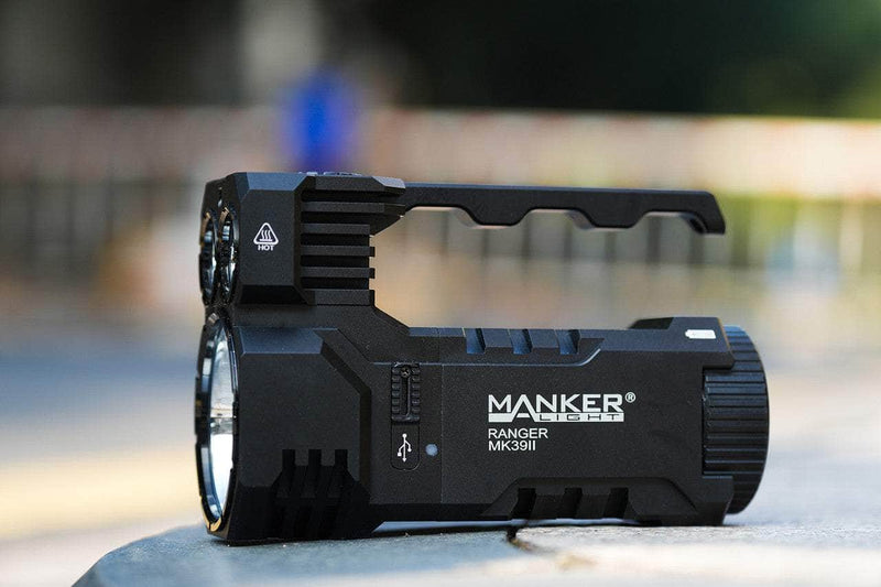 A black Manker Ranger MK39 II flashlight with a handle rests on a flat surface outdoors, its unique design perfect for spotlight and long-range use against the blurred background.