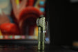 Close-up of a Manker E02 III AAA / 10440 Flashlight, standing upright and providing 560 lumens of illumination in green. The blurred background features colorful objects that accentuate this brightest everyday carry angle flashlight with its best-in-class features.