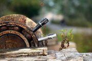Pair of Manker E02 III AAA / 10440 Flashlights with high-performance LED lights illuminate a rustic wooden wheel, surrounded by greenery and a softly blurred background.