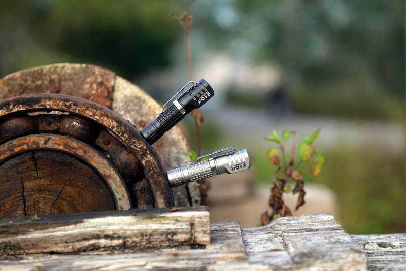 Pair of Manker E02 III AAA / 10440 Flashlights with high-performance LED lights illuminate a rustic wooden wheel, surrounded by greenery and a softly blurred background.