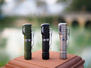 Three Manker E02 III flashlights in green, black, and silver stand upright on a wooden surface outdoors. Equipped with high-performance LED technology, they deliver 560 lumens of illumination. Perfect for the brightest everyday carry needs, they shine against the blurred greenery backdrop.