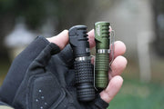 A gloved hand holds two small, textured Manker E02 III AAA / 10440 flashlights. One is black and the other is green, featuring high-performance LED technology. These are the brightest everyday carry tools with up to 560 Lumens of illumination.