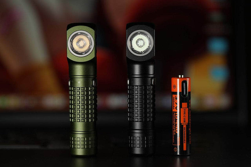 Two Manker E02 III flashlights, one in olive green and the other in black, stand proudly on a surface next to an AA battery, showcasing their high-performance LED technology. With an impressive 560 lumens illumination, they promise to be the brightest everyday carry in any situation. The background remains softly blurred.