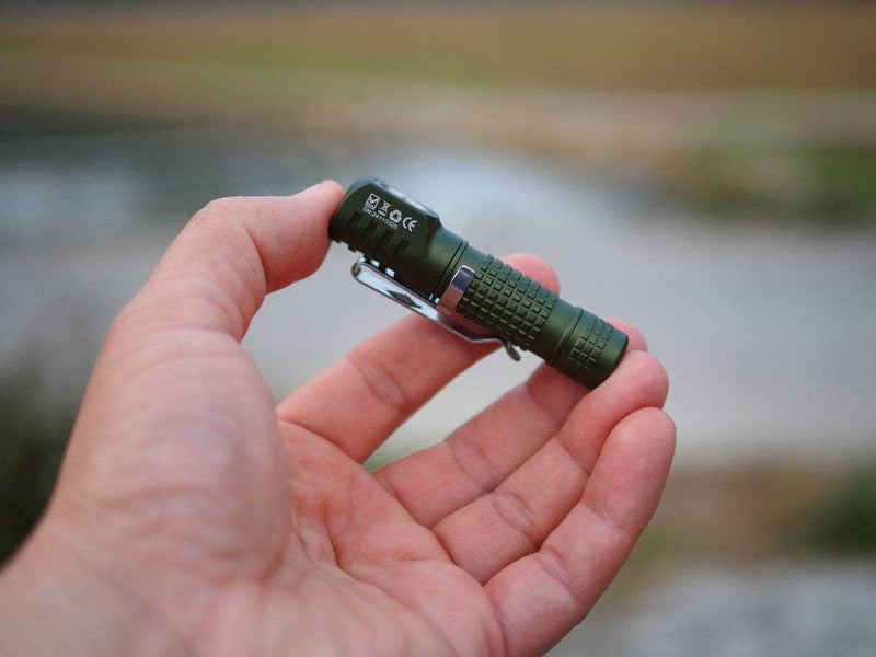 A Manker E02 III AAA / 10440 flashlight in green, showcasing its best-in-class features, is held in a hand outdoors against a blurred background.