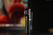 The Manker E02 III AAA / 10440 flashlight, in its sleek black design, stands upright on a dark surface. Its best-in-class features are highlighted against a blurred colorful background while delivering an impressive illumination of 560 lumens for any setting.