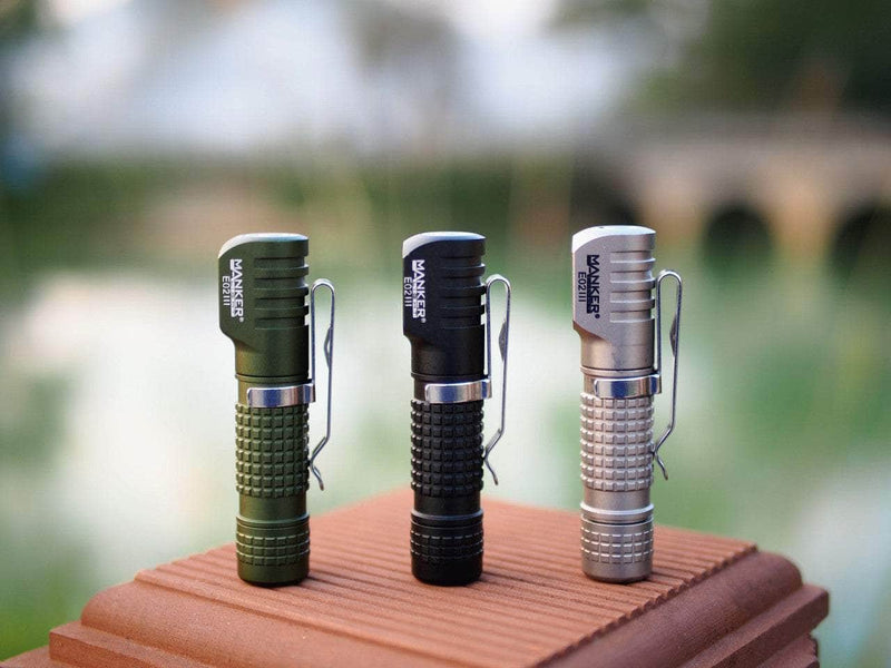 Three Manker E02 III AAA / 10440 flashlights in green, black, and silver stand upright on a brown surface with a blurred outdoor background, demonstrating their high-performance LED capabilities. Perfect for the brightest everyday carry needs, each offers 560 lumens of illumination.