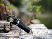 The Manker E02 III AAA / 10440 Flashlight, featuring a black finish and textured grip with high-performance LED, is resting at an angle on a weathered wooden surface outdoors.
