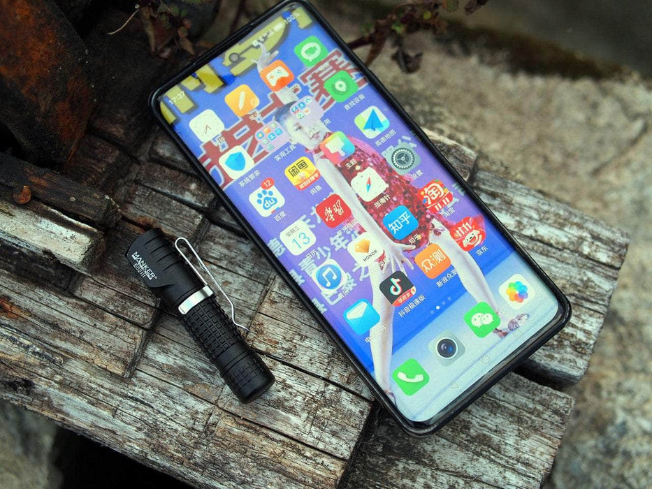 A smartphone with a colorful app-filled screen sits on a wooden surface next to the Manker E02 III AAA / 10440 Flashlight, which boasts high-performance LED technology and offers 560 lumens illumination for the brightest everyday carry.