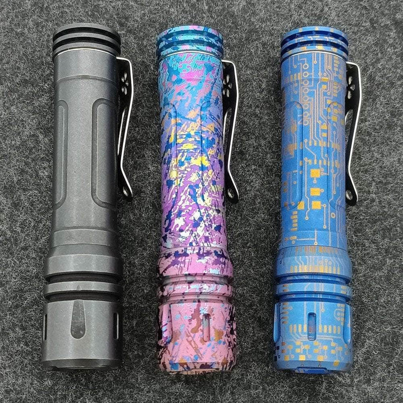 Three uniquely designed flashlights stand upright on a gray textured surface. Featuring matte black, colorful abstract, and blue circuit board patterns, each has a metal clip. One is a ReyLight Anodized Ti LAN flashlight with Nichia 519A Hi-CRI LED for superior illumination.