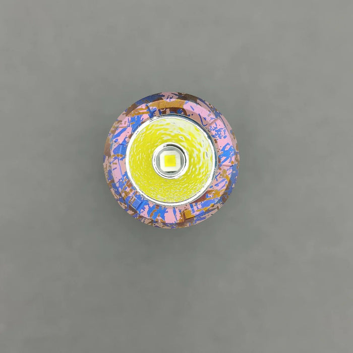 A top-down view of a colorful LED flashlight with the XPL HI 6000K LED and a patterned pink and blue surface, reminiscent of the ReyLight Anodized Ti LANapple, against a plain gray background.