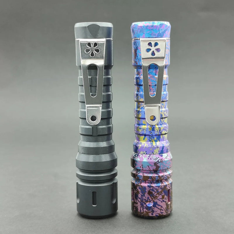 Two flashlights stand against a plain backdrop. The left is sleek black titanium, and the right one showcases a colorful splattered design like the ReyLight Anodized Ti LANapple. Both have metal clips with flower-like cutouts and are equipped with XPL HI 6000K LEDs.