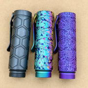 Three cylindrical metal flashlights—including the ReyLight Dawn Hex - Ti Anodized, a multicolored model, and a purple design with swirls—stand vertically on a tan surface.