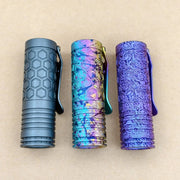 Three compact flashlights—one being the ReyLight Dawn Hex - Ti Anodized—feature black hexagonal, multicolored swirled, and purple patterned finishes. All stand upright on a beige surface.