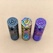 Three cylindrical metal flashlight bodies, including the ReyLight Dawn Hex - Ti Anodized—black, gold with rainbow accents, and purple—stand upright on a plain beige surface.