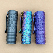 Three pocket flashlights, including the ReyLight Dawn Hex - Anodized, display unique textures and colors—black with hexagons, iridescent blue, and purple—standing upright on a beige surface.