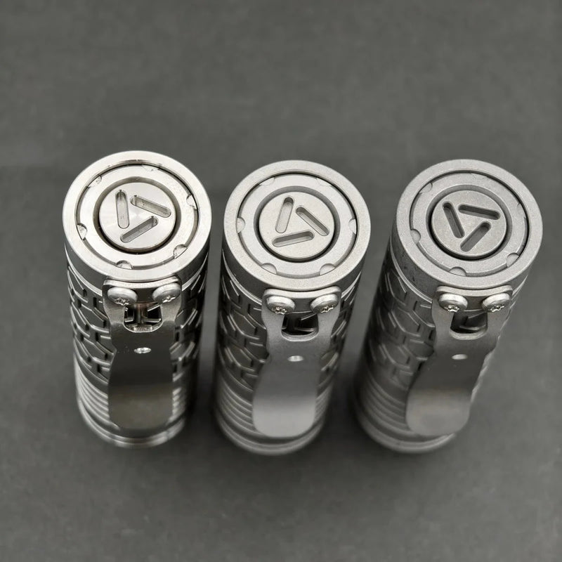 Three ReyLight Dawn Hex - Ti flashlights with textured titanium bodies and pocket clips, powered by rechargeable 18350 batteries and equipped with Nichia 519a LEDs, are arranged side by side on a dark gray surface, viewed from above.