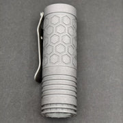 A compact ReyLight Dawn Hex - Ti flashlight with a hexagonal pattern and metal clip rests on a dark surface, powered by a rechargeable 18350 battery.
