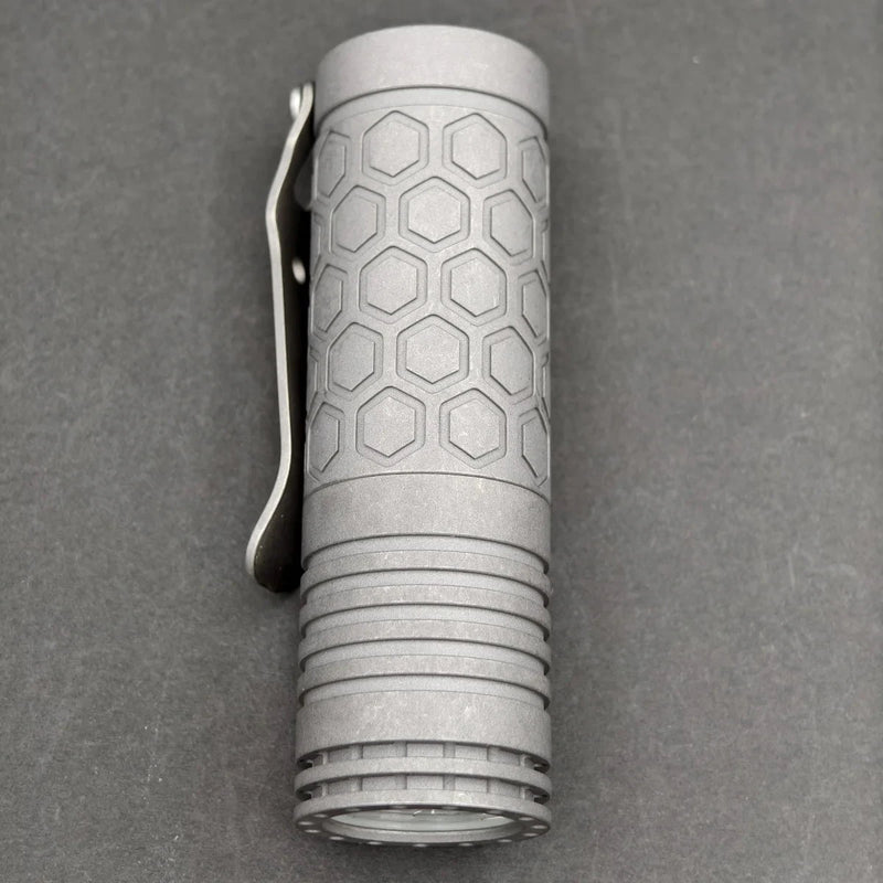 A compact ReyLight Dawn Hex - Ti flashlight with a hexagonal pattern and metal clip rests on a dark surface, powered by a rechargeable 18350 battery.
