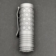 A compact ReyLight Dawn Hex - Ti flashlight with a hexagonal pattern, metal clip, and Nichia 519a LED rests on a dark gray surface.