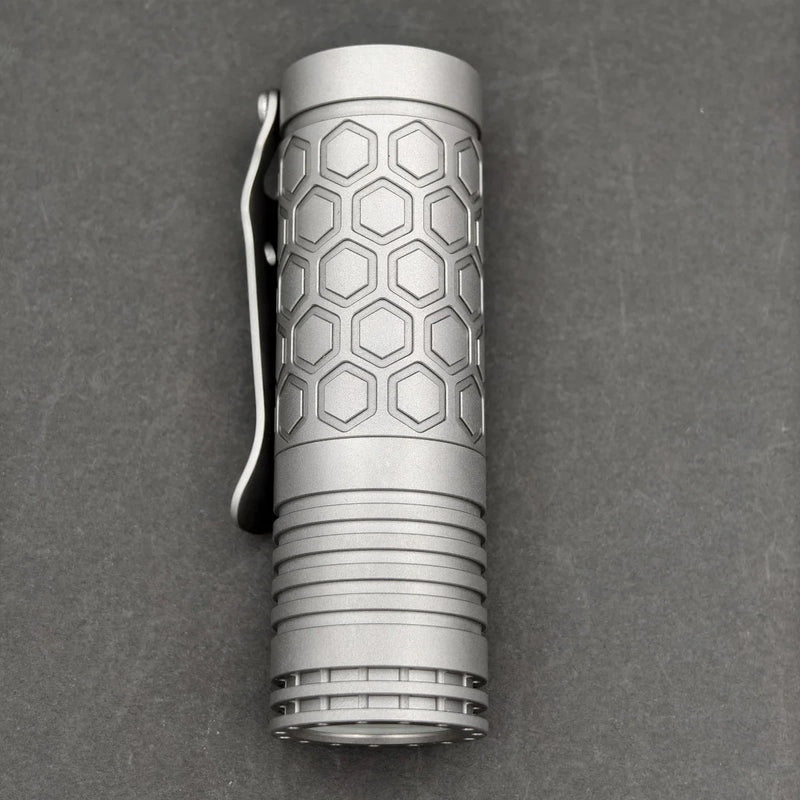 A compact ReyLight Dawn Hex - Ti flashlight with a hexagonal pattern, metal clip, and Nichia 519a LED rests on a dark gray surface.