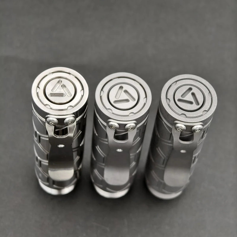 Three ReyLight Dawn Hex - Ti flashlights stand upright on a grey surface, each with a triangular logo on the tail cap, a metal pocket clip, and powered by rechargeable 18350 Li-ion batteries.