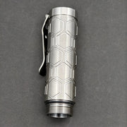 The ReyLight Dawn Hex - Ti is a titanium flashlight tube with a hexagonal pattern and pocket clip, shown on a dark background. It features a Nichia 519a LED and runs on a rechargeable 18350 Li-ion battery.