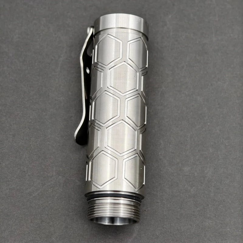 The ReyLight Dawn Hex - Ti is a titanium flashlight tube with a hexagonal pattern and pocket clip, shown on a dark background. It features a Nichia 519a LED and runs on a rechargeable 18350 Li-ion battery.