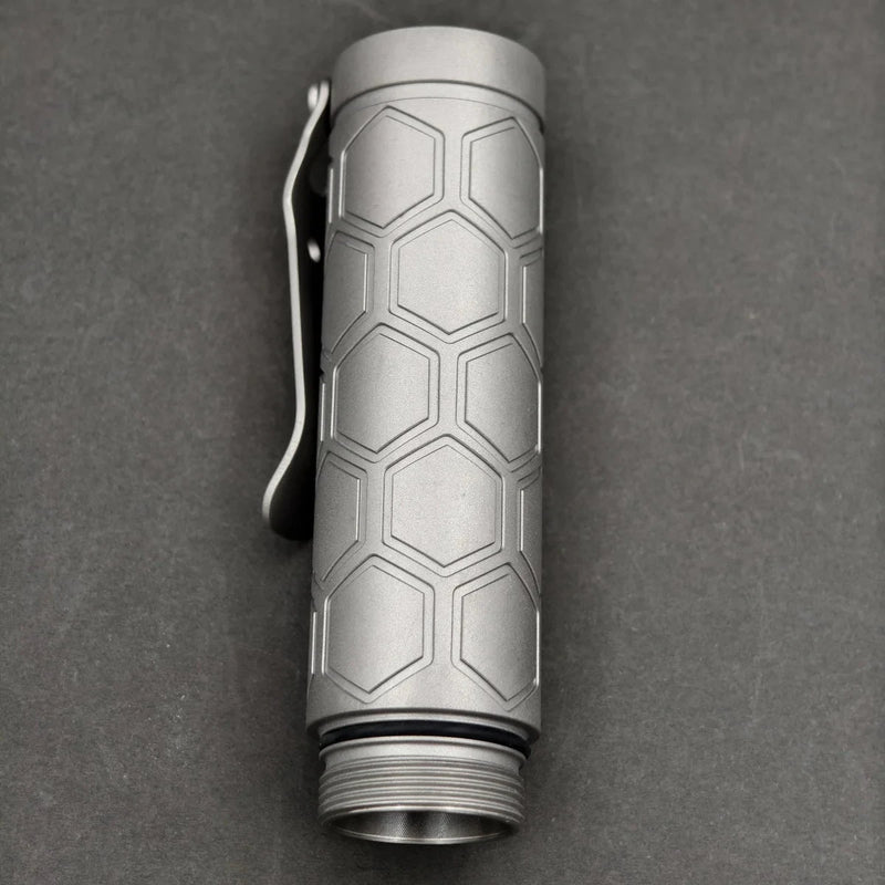 A ReyLight Dawn Hex - Ti flashlight featuring a hexagonal patterned grip, pocket clip, and Nichia 519a LED is displayed against a plain dark background.