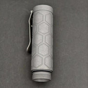 The ReyLight Dawn Hex - Ti is a cylindrical flashlight with a hexagonal pattern, pocket clip, Nichia 519a LED, and threaded end cap, shown on a dark surface.