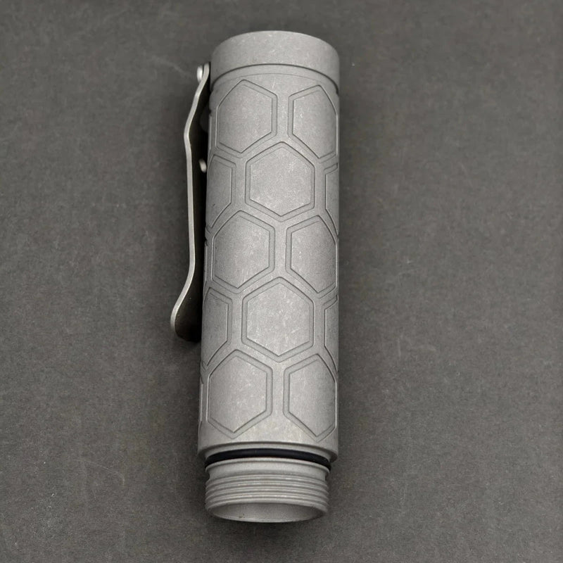 The ReyLight Dawn Hex - Ti is a cylindrical flashlight with a hexagonal pattern, pocket clip, Nichia 519a LED, and threaded end cap, shown on a dark surface.