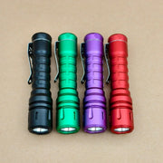 Four compact ReyLight LANapple - Aluminum flashlights in black, green, purple, and red are side by side on a brown surface. Each uses a Nichia 519a 4000K LED and is powered by a USB-C rechargeable 14500 battery.